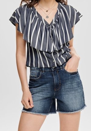 Vrouw die een navyblauw-wit geruite blouse met ruches draagt en een donkere, hoog opgesneden denimshort met opgerolde zoom.