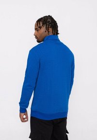 Pull zippé en tricot bleu avec un motif texturé et des poignets côtelés. Modèle vu de dos, le portant avec un pantalon cargo noir.