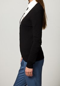 Svart kofta i en figurnära stil med långa ärmar, tillverkad av mjukt material, tillsammans med en vit skjorta med krage och blå byxor.