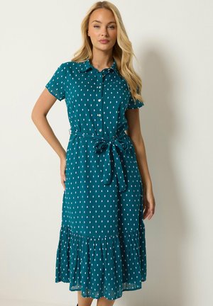 Blonde Frau trägt ein türkisfarbenes Midikleid mit weißen rechteckigen Punkten, kurzen Ärmeln, Knöpfen vorne, gebundenem Taillengürtel und Rüschen am Saum.