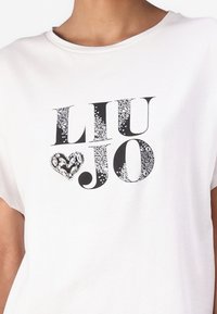 Balta trumpa rankovė T-marškiniai su "LIU JO" juodu šriftu, papuošti tekstūruotu širdies dizainu, užpildytu sidabro akcentais.