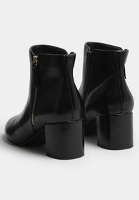 Botas negras hasta el tobillo con tacones cuadrados y cremalleras laterales, vistas desde atrás sobre un fondo blanco liso.