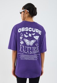 Camiseta de gran tamaño en color púrpura con un diseño gráfico blanco que incluye mariposas y elementos de texto en la parte trasera. Fabricada en algodón, con corte holgado.