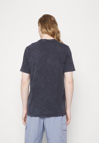 Person with long hair facing away, wearing a dark washed short-sleeve t-shirt and light blue cargo pants against a plain background.