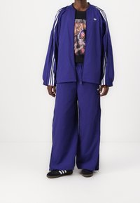 Chándal morado con rayas blancas, que incluye una chaqueta con cremallera y pantalones con cintura elástica, combinado con sandalias negras y una camiseta gráfica.