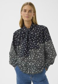 Woman wearing a long-sleeve floral blouse with a large bow tie at the neck, paired with blue jeans, standing with hands in pockets.