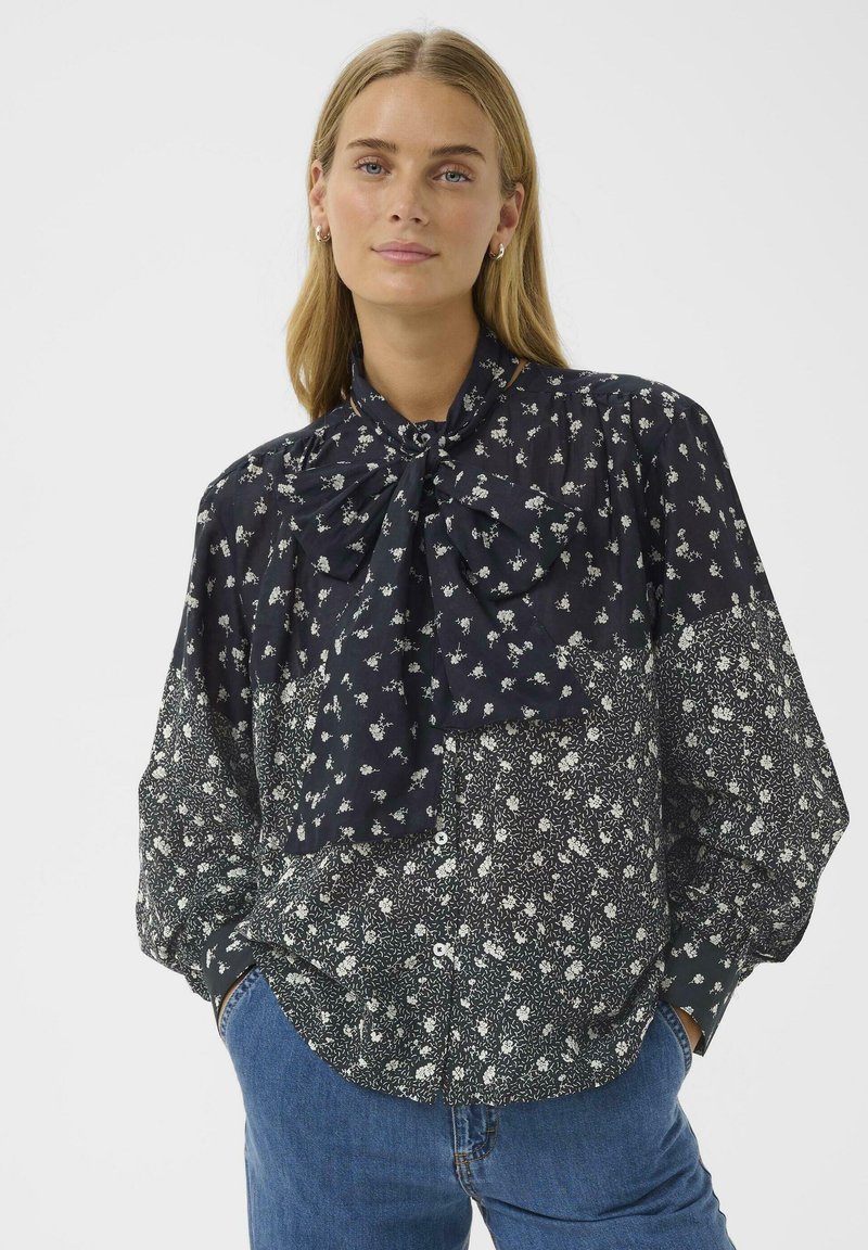 Woman wearing a long-sleeve floral blouse with a large bow tie at the neck, paired with blue jeans, standing with hands in pockets.