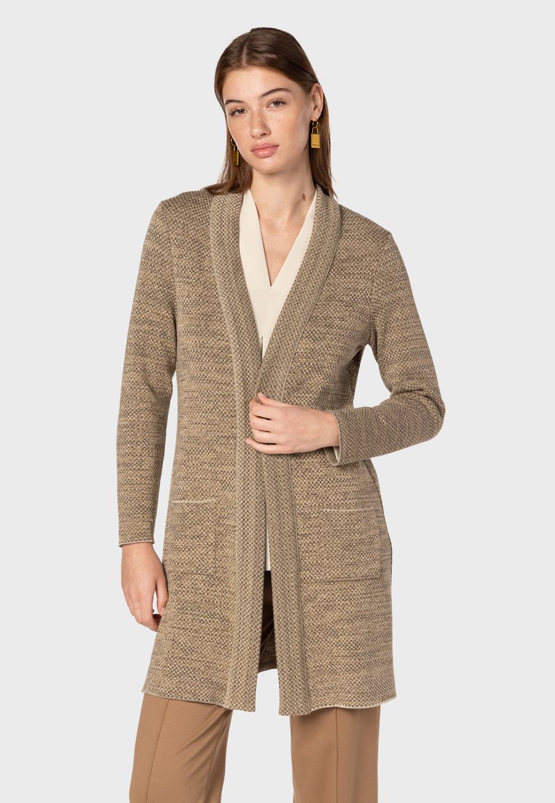 Lång cardigan i beige stickat med ett strukturerat mönster, öppen fram, två framfickor och en sjalkrage. Tillsammans med en ljus topp och byxor.