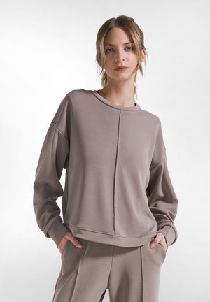 Jonge vrouw met haar in een staart, draagt een losse taupekleurige trui en bijpassende broek met handen in de zakken tegen een witte achtergrond.
