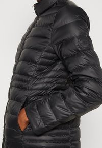 Veste noire matelassée avec texture en losanges, col montant et poches zippées. Présente un logo cousu sur la manche gauche.