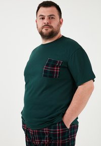 Vihreä lyhythihainen t-paita, jossa on ruutukuviollinen taskuyksityiskohta. T-paidassa on pyöreä pääntie ja rento istuvuus, yhdistettynä ruutukuosisisiin aamutohveleihin.