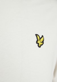 Camiseta blanca de algodón con un pequeño logotipo de pájaro bordado en negro y amarillo en el lado izquierdo. Textura suave, diseño sencillo.