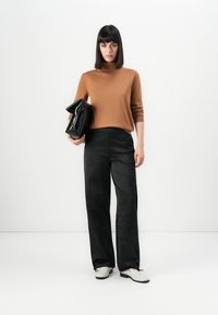 Vrouw met steil donker haar draagt een bruine coltrui, zwarte brede broek, witte schoenen en houdt een grote zwarte clutch tas vast, staand tegen een witte achtergrond.