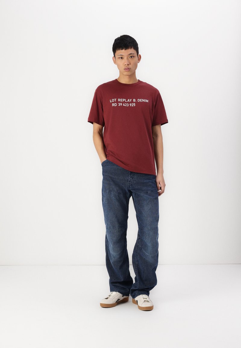Bordeauxfarbenes T-Shirt mit Rundhalsausschnitt und weißem Text, kombiniert mit blauen Jeans aus Denim, die vertikale Streifen und gestickte Akzente aufweisen. Weiße Sneaker vervollständigen den Look.