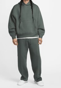 Tmavě zelená oversized mikina s kapsou, doplněná o odpovídající tapered tepláky. Hladká tkanina, uvolněný střih, bílé akcenty na pásku.