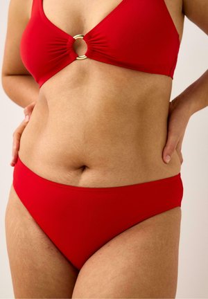 Kvinde iført en rød todelt badedragt med en guldringdetalje på toppen, hænderne hvilende på hofterne, synlig overkrop og øvre ben.