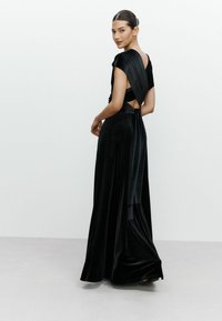 Rochie din catifea neagră, având un design drapat, cu mâneci scurte și spate descoperit. Fusta coboară până la podea, creând o siluetă elegantă.
