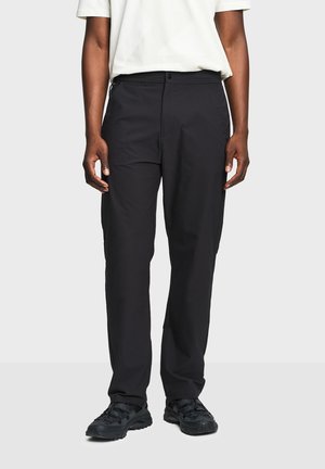 Osoba u ravnim crnim hlačama, crnim tenisicama i bijeloj majici kratkih rukava, stoji na jednobojnoj svijetloj pozadini.