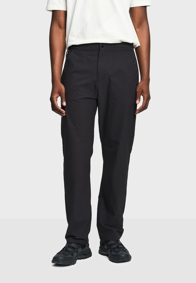 Άτομο που φοράει μαύρο παντελόνι με ίσια γραμμή, μαύρα αθλητικά παπούτσια και λευκό κοντομάνικο πουκάμισο, στέκεται σε απλό φωτεινό φόντο.