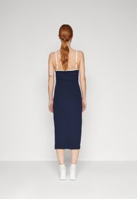 Žena s dlouhými rudými vlasy v nízkém ohonu, oblečená v námořnickomodré bezrukávové midi šaty a bílé tenisky, stojící proti jednobarevnému šedému pozadí.