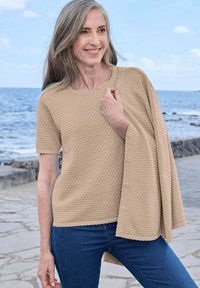 Gl smiling vrouw met lang grijs haar, gekleed in een beige textuur kortemouwen top en een bijpassende knopen cardigan vasthoudend aan zee.
