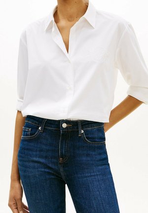 Femme portant une chemise blanche à boutons avec les manches retroussées et un jean bleu foncé taille haute, debout devant un fond uni.