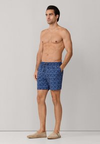 Homme torse nu portant un short de bain bleu à motifs et des chaussures beige à enfiler, debout, les mains dans les poches, sur un fond uni.