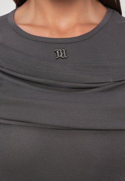 Zbliżenie na osobę w szarym, prążkowanym topie z zakładanym dekoltem i małą, czarną haftowaną literą "M" na środku klatki piersiowej.
