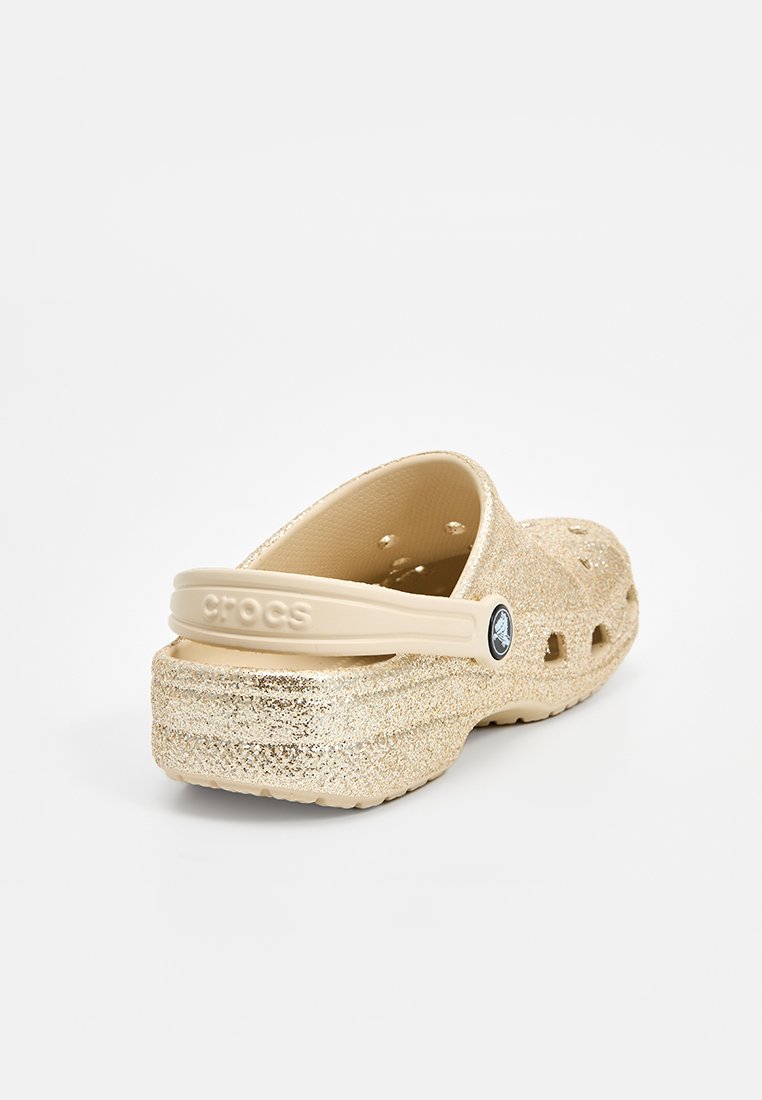 Des Crocs dorées pailletées avec une bride à l'arrière, fabriquées en matériau léger, dotées de trous de ventilation, d'une semelle texturée et du logo de la marque sur le côté.