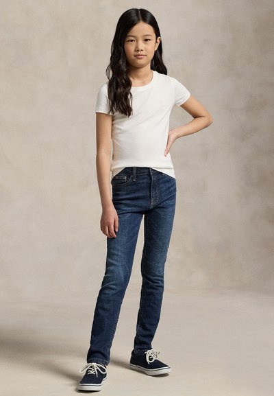 Jeune fille aux longs cheveux foncés portant un t-shirt blanc à manches courtes, un jean bleu et des baskets bleu marine, debout avec une main sur la hanche.