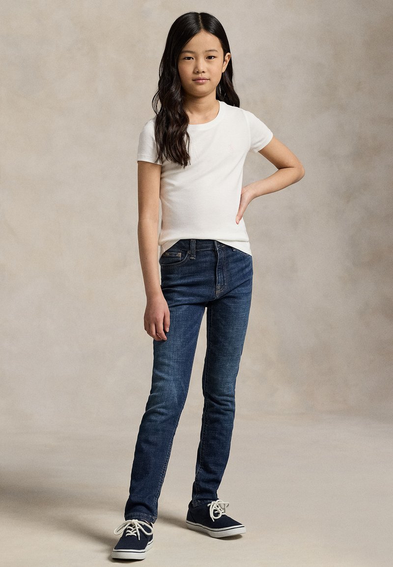 Jeune fille aux longs cheveux foncés portant un t-shirt blanc à manches courtes, un jean bleu et des baskets bleu marine, debout avec une main sur la hanche.