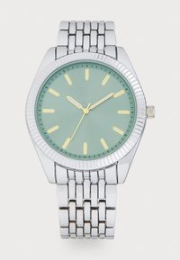 Montre en métal argenté avec un boîtier arrondi, un cadran vert menthe, des index horaires jaunes et une lunette texturée. Elle possède un bracelet en métal à maillons.