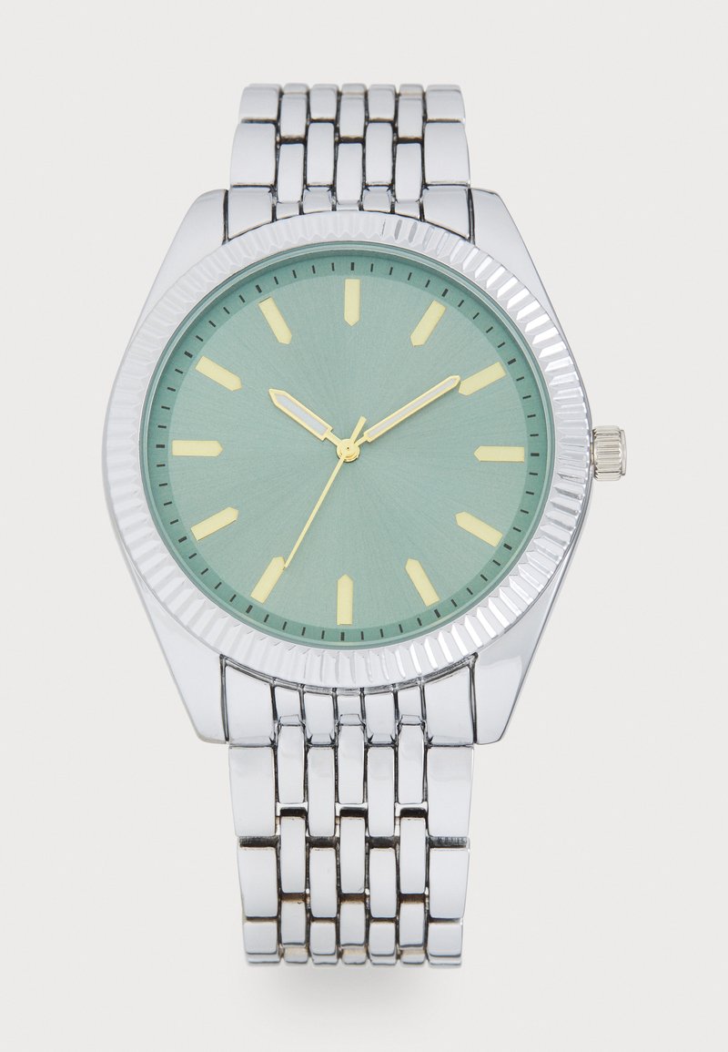 Montre en métal argenté avec un boîtier arrondi, un cadran vert menthe, des index horaires jaunes et une lunette texturée. Elle possède un bracelet en métal à maillons.