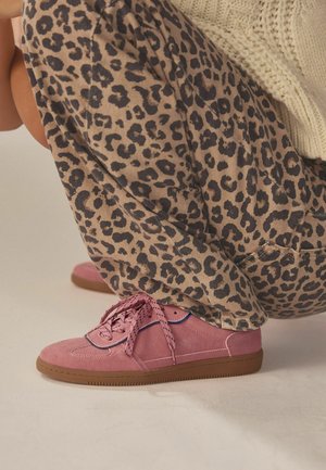 Pink suede sneakers with textured laces and a brown rubber sole, paired with beige pants featuring a black leopard print.