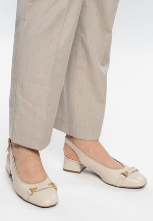 Chaussures beige à talon bloc et bride arrière avec détails en boucle dorée, portées avec un pantalon large beige clair, sur fond blanc.