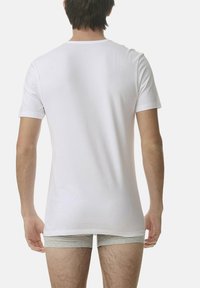Vit t-shirt med kort ärm och rund halsringning, tillverkad av ett glatt tyg. Bakifrån visas en avslappnad passform, i kombination med ljusgrå kalsonger.