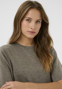 Pelēka īsām piedurknēm t-krekls ar apaļu dekoltē. Mīksta, tekstūrēta auduma kontūras maigi pieguļ. Modelim ir viļņaini mati un viņš pozē ar saliktām rokām.