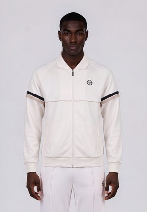 Homme portant une veste blanche zippée avec des rayures noires et beiges sur les manches et un pantalon blanc assorti avec un logo sur la poitrine et la cuisse.
