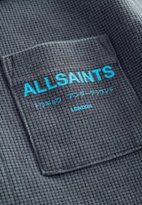 Tissu gris foncé texturé avec une poche affichant un texte bleu vif « ALLSAINTS » et des mots plus petits en japonais et en anglais en dessous.