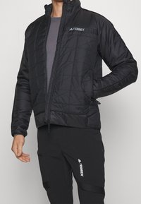 Czarna pikowana kurtka z wysokim kołnierzem, z zamkiem błyskawicznym z przodu i logo TERREX na klatce piersiowej. Gładka tekstura i lekka konstrukcja.