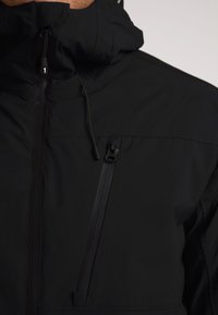 Giacca nera impermeabile con colletto alto, zip frontale, cappuccio regolabile e tasca sul petto con zip. Tessuto liscio con finitura opaca.