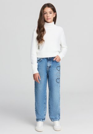 Maglione bianco a coste spessa con collo alto abbinato a jeans a gamba larga di colore blu chiaro, con decorazioni a forma di cuore in argento sui lati.