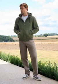 Olivgrüner Zip-Hoodie mit gefütterter Kapuze, kombiniert mit hellbraunen Hosen und grauen Sneakers mit weißen Akzenten.