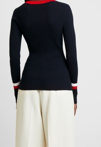 Osoba oblečená v přiléhavém námořnickém žebrovaném svetru s červenými a bílými pruhy na límci a manžetách, v kombinaci s širokými krémovými kalhotami.