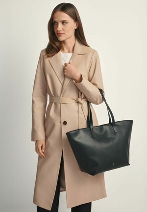 Kobieta w beżowym płaszczu trencz z paskiem, trzymająca dużą czarną skórzaną torbę na ramię, stojąca na jednolitym tle.