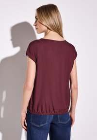 Blouse courte bordeaux à manches courtes avec une coupe ample et un ourlet élastique. Texture douce, encolure ronde et manches retroussées. Portée avec un jean bleu.