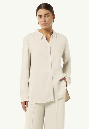Jasno beżowa, długosleeve koszula z guzikami, wykonana z miękkiego materiału, z kołnierzykiem i luźnym krojem. Model ma jedną rękę w kieszeni.