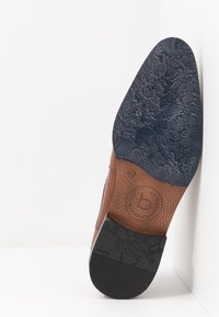 Semelle de chaussure en cuir marron avec un fond en caoutchouc noir texturé, présentant un logo circulaire et des motifs de surface détaillés.