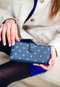 Navy blue wallet made of textured material, featuring light blue polka dots and a circular black clasp with a logo.