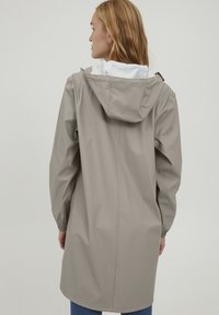 Beige nepromokavá bunda s kapucí, která má elastické manžety a volný střih. Vnitřek je s kontrastní bílou podšívkou.